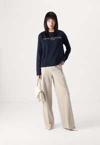 Tengerészkék hosszú ujjú póló "TOMMY HILFIGER" logóval, bézs széles szárú nadrággal és fehér kézitáskával, láncos pánttal. Egyszerű, modern dizájn.