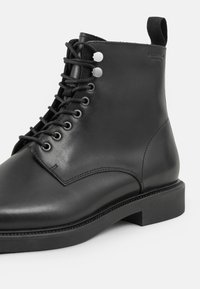 Crne kožne gležnjače s glatkom završnom obradom, okruglim prstom, vezanjem sprijeda i malom petom. Ima srebrne rupice za vezanje i zadnji čep za nošenje.