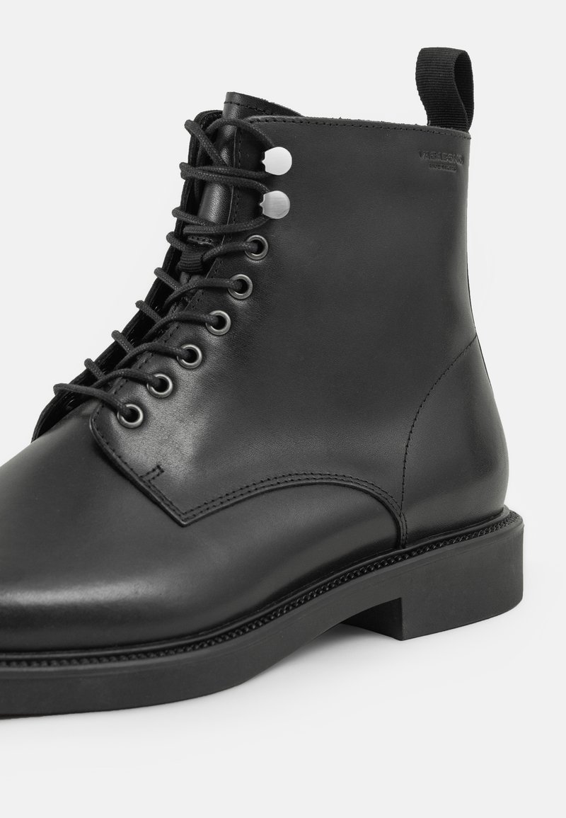 Zwarte leren enkelboot met een gladde afwerking, ronde neus, vetersluiting aan de voorkant en een kleine hak. Bevat zilveren oogjes en een lus aan de achterkant.