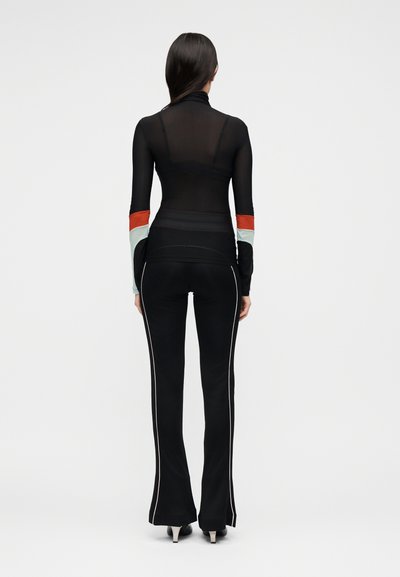 Γυναίκα που φοράει μαύρη διάφανη μπλούζα με μακριά μανίκια με ρίγες κόκκινου και ανοιχτού μπλε χρώματος στα μανίκια, μαύρο φαρδύ παντελόνι με λευκές πλαϊνές ρίγες και ψηλοτάκουνα, στέκεται με την πλάτη.