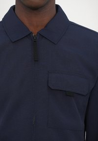 Navy blue jacket made of lightweight fabric, featuring a zipper closure and a patch pocket on the left side. Solid color with a subtle texture.