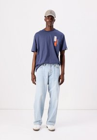 Navy blue cotton t-shirt with a chest graphic, paired with light blue loose-fitting jeans and beige sneakers. Model wears a gray cap.