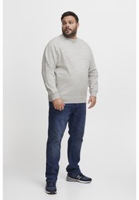 Man in een lichtgrijze trui, donkerblauwe jeans en zwarte sneakers, staande tegen een witte achtergrond, kijkend naar links.