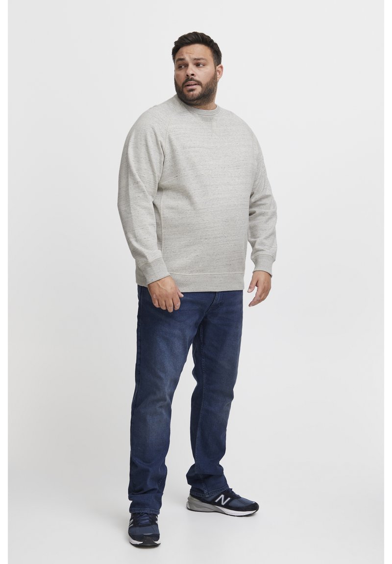 Man in een lichtgrijze trui, donkerblauwe jeans en zwarte sneakers, staande tegen een witte achtergrond, kijkend naar links.