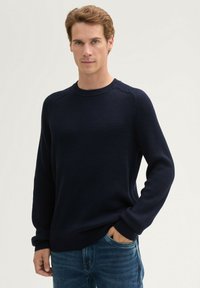 Pull en maille côtelée marine avec col rond et manches longues, associé à un jean bleu. Présente une texture horizontale et une coupe décontractée.