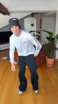 Langermet hvit skjorte med grafikk og tekst på ermet, svart bøttehatt, svarte vide bukser, og sølvfargede joggesko. Innendørs setting med planter.