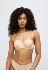 Beige polstret BH med tynne justerbare stropper, glatt stoff, subtil blondekant og en liten sløyfedetalj i midten. Ensfarget bakgrunn.