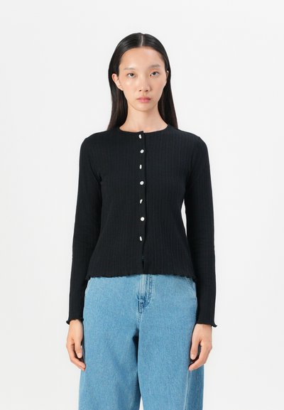 Musta ribbed pitkähiha cardigan, jossa on pyöreä kaula-aukko. Edessä on kuusi valkoista nappia ja se on lyhyt, laineileva helma.