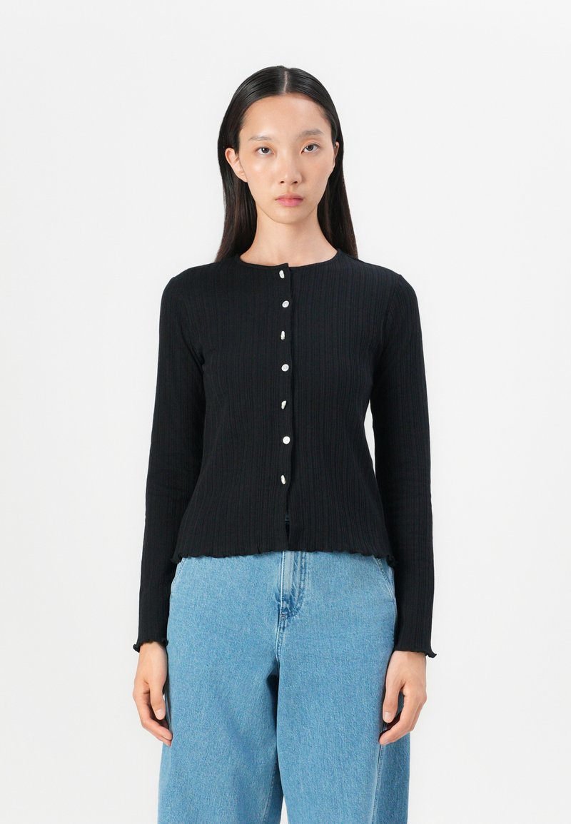 Black ribbed long-sleeve cardigan with a rounded neckline, featuring six white buttons down the front and a cropped, scalloped hem.