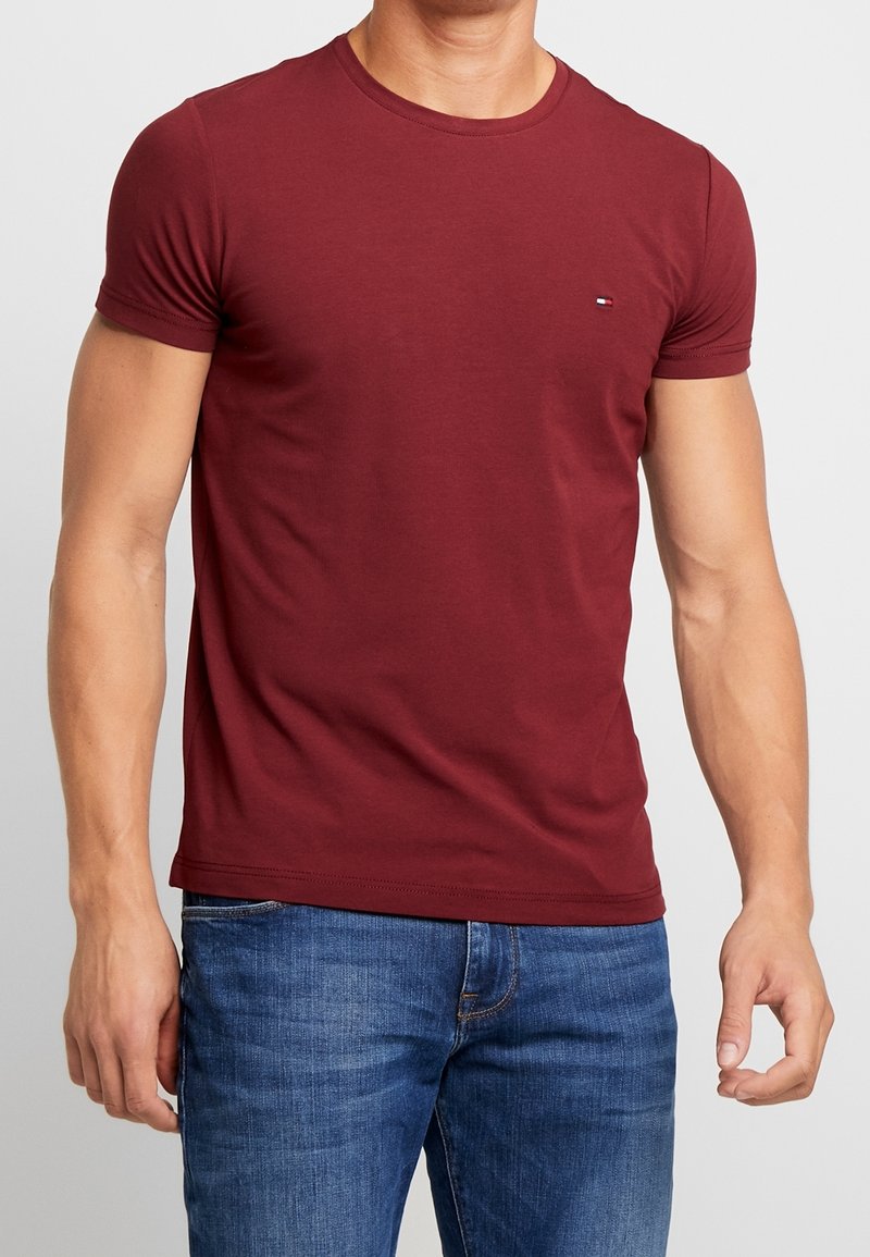 Camiseta de algodón granate con un cuello redondo clásico y mangas cortas, con un pequeño logotipo en el pecho. Combinada con vaqueros azules.