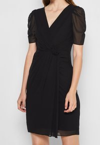 Černé šaty s výstřihem do V, krátkými nafouklými rukávy a skládáním v pase s detaily v podobě růže. Hladká látka s volným střihem.