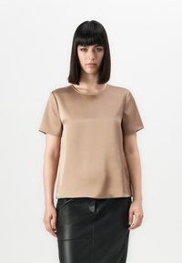 Vrouw met steil donker haar, gekleed in een beige satijnen top met korte mouwen en een zwarte leren rok, staand tegen een effen witte achtergrond.