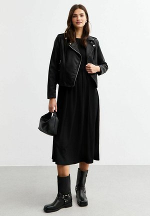Veste de motard en cuir noir portée sur une robe maxi noire. Bottines noires avec ferrures en argent. Petit sac à main noir. Texture lisse, silhouette ajustée.