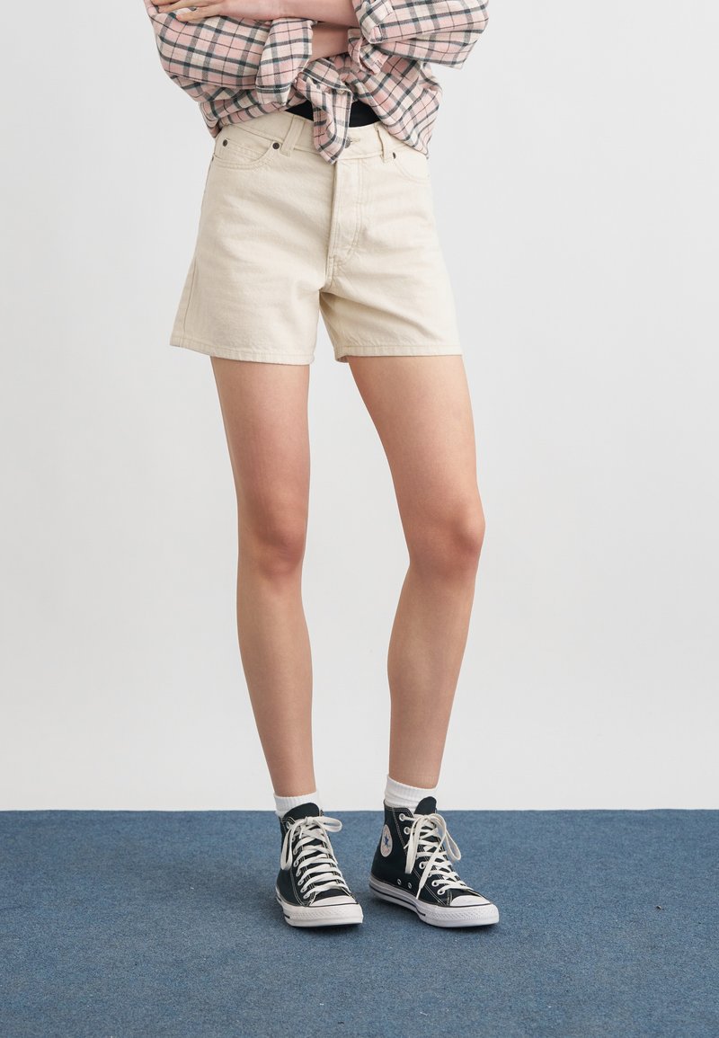 Personne portant un short en denim beige, des baskets montantes noires avec des lacets blancs, des chaussettes blanches et une chemise à carreaux retroussée, les bras croisés.