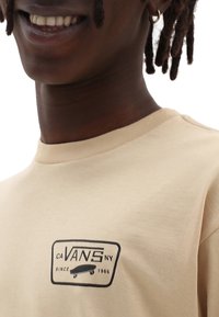 Lächelnde Person mit beigem Vans-T-Shirt mit schwarzem Logo und Skateboardgrafik, goldenen Creolen und kurzen Dreadlocks.