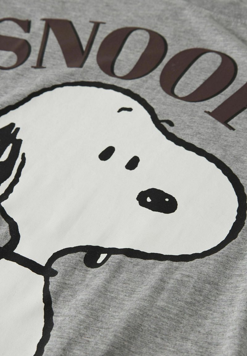 Το πρόσωπο του χαρακτήρα Snoopy σε ασπρόμαυρο πάνω σε γκρι ύφασμα με μερικό κείμενο "SNOOP" πάνω από το κεφάλι, πιθανό μέρος ενός σχεδίου μπλουζάκι ή φούτερ.