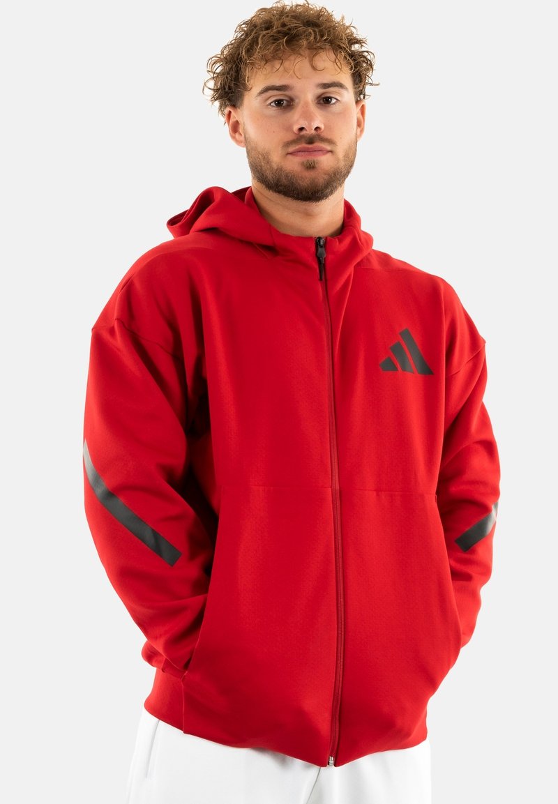 Felpa rossa con zip e cappuccio, con dettagli neri sulle maniche e un logo sul petto. Tessuto morbido con una vestibilità rilassata.