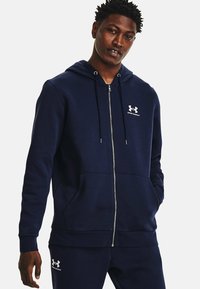 Jūras zilas pievilkšanas kapuces džemperis ar mīkstu tekstūru un baltu Under Armour logo uz krūtīm. Iespējas ietver priekšējās kabatas un ribotās piedurknes.