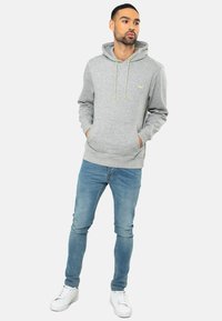 Grijze hoodie met een trekkoord en voorzakken, gedragen met lichtblauwe jeans en witte sneakers. Bevat een groen logo op de borst.