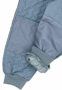 Detail am Ärmel einer blauen Steppjacke mit gerippten Bündchen und abgegrenzten Stoffpaneelen, die Textur und Steppmuster zeigen.