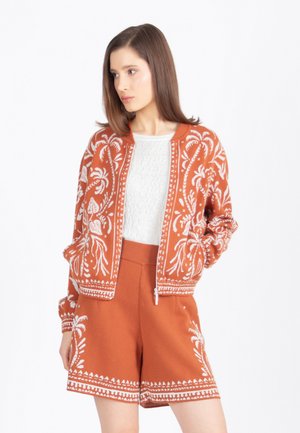 Noor naine kannab roostepunast mustrilist jopet ja sobivaid lühikesi pükse, valge topp seljas, vaatab valgele taustale külili.