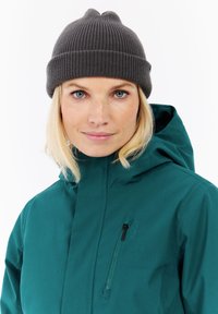 Mujer con cabello rubio claro que lleva un gorro de punto gris oscuro y una chaqueta verde azulado con un bolsillo con cremallera en el pecho, mirando hacia adelante con una ligera sonrisa.
