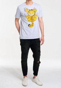 Camiseta de algodón gris con un gráfico de un gato de dibujos animados amarillo con rayas negras. Combinada con jeans negros desgastados y zapatillas blancas.