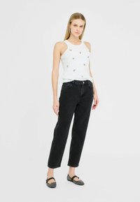 Giovane donna che indossa un top bianco senza maniche con decorazioni, jeans neri a vita alta e ballerine nere con motivo, in piedi su uno sfondo bianco.
