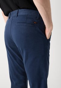 Pantaloni bleumarin dintr-un material neted, cu o croială slim, buzunare la spate și un mic etichetă portocalie cu logo-ul "BOSS" pe talie.