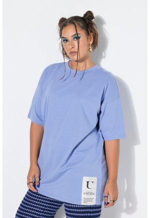 Femme avec deux mèches frontales tressées et des boucles d'oreilles créoles dorées porte un t-shirt ample bleu clair et des leggings bleu marine à motifs.