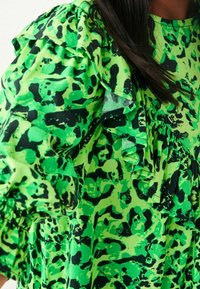 Robe abstraite à motifs verts et noirs avec des détails froncés aux épaules et aux manches, portée par une personne aux cheveux foncés.