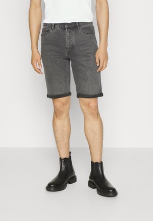 Personne portant un short en jean noir retroussé, un t-shirt blanc et des bottines en cuir noir, debout devant un fond blanc.