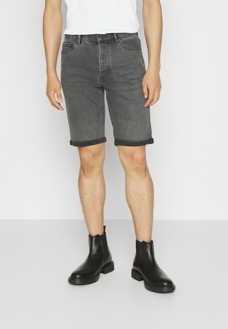 Personne portant un short en jean noir retroussé, un t-shirt blanc et des bottines en cuir noir, debout devant un fond blanc.