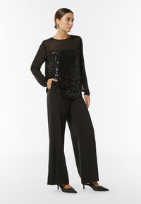 Schwarzes, transparentes Langarmoberteil mit einem Paillettenmuster an der Vorderseite, kombiniert mit hochtaillierten, schwarzen weiten Hosen und spitzen, schwarzen High Heels.