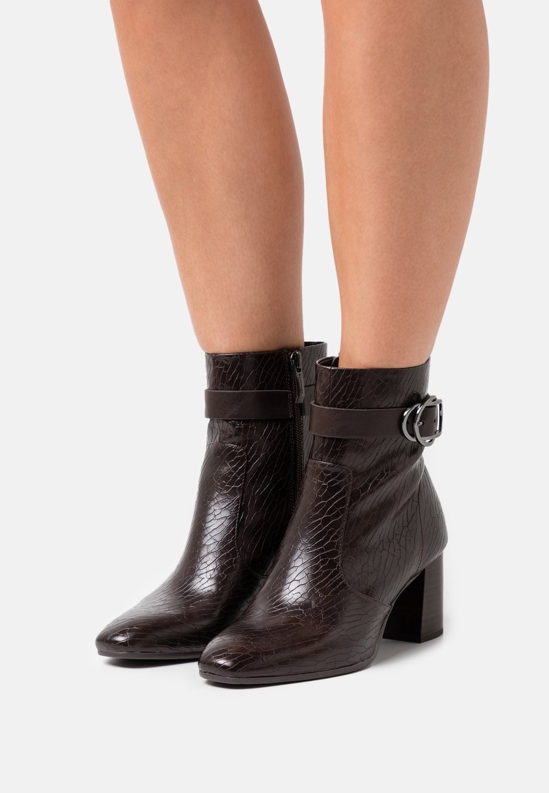 Botas de cuero de color marrón oscuro, con relieve de cocodrilo, que llegan hasta los tobillos. Tienen punteras puntiagudas, Tacones de bloque, cremalleras en los laterales y correas decorativas con hebillas plateadas.
