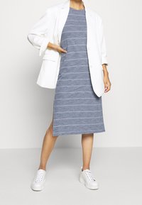 Robe midi à rayures bleues en tissu léger, associée à un blazer blanc oversize. Elle présente un col rond et des fentes sur les côtés; portée avec des baskets blanches.