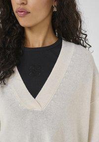 Pull-over crème à col en V avec une coupe ample, présentant des bords côtelés et une texture subtile. Porté par-dessus une chemise noire à manches longues côtelée.
