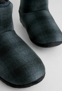 Papuci pufoși verde-albăstrui, cu o textură moale, vârful rotund și o talpă din cauciuc negru. Include un model subtil pe material.