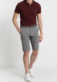 Polo bourgogne avec trois boutons, shorts gris avec un motif texturé, ceinture marron et baskets blanches sur un fond uni.