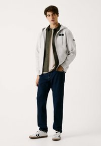 Sudadera gris claro con cordones, superpuesta a una camiseta verde oscura, combinada con una camiseta beige y jeans azul oscuro. Zapatillas blancas con acentos negros.
