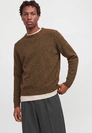 Homme portant un pull en maille marron sur une chemise beige et un pantalon ample gris foncé, debout, ajustant son pull d'une main.