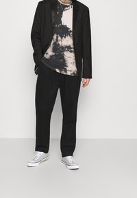 Chaqueta de traje negra sobre una camiseta de tie-dye en tonos de negro y rosa pálido, combinada con pantalones negros y zapatillas grises claras.
