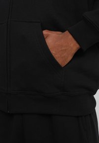 Sudadera negra con un bolsillo frontal, puños y cinturilla acanalados. El material parece suave con una textura blanda. Mano descansando en el bolsillo.