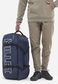 Grand sac de sport navy, fabriqué en matériau durable, avec des sangles noires et un grand texte blanc. Personne en pantalon beige et chaussures noires à côté.