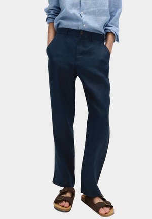 Persona con pantalones azul marino, camisa de manga larga azul claro y sandalias marrones de doble correa, de pie con las manos en los bolsillos.