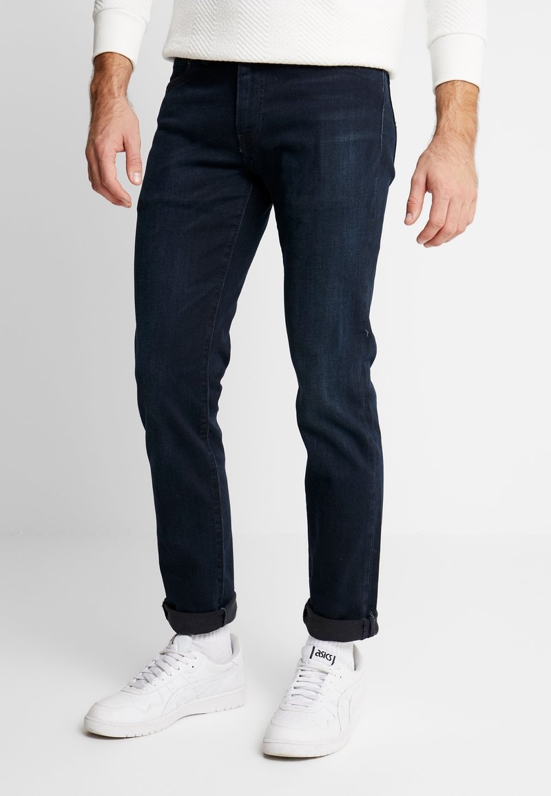 Homme portant un jean slim bleu foncé avec les revers retroussés, des baskets blanches Asics et une chemise blanche texturée à manches longues.