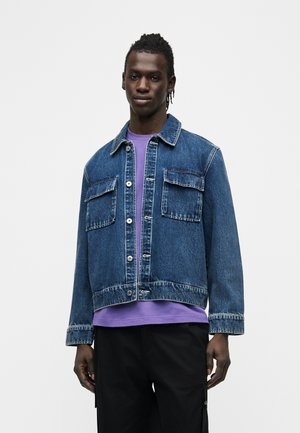 Uomo che indossa una giacca di denim blu sopra una camicia viola e pantaloni neri, in piedi davanti a uno sfondo bianco uniforme.