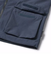Mörkblå tygjacka med zipplock fram, en praktisk ficka och kardborrestängning. Texturen har ett subtilt rutmönster.
