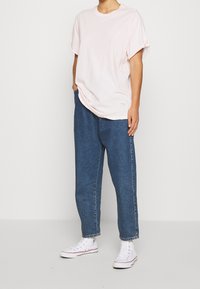 Jasnoróżowa bawełniana koszulka z podwiniętymi rękawami, zestawiona z luźnymi niebieskimi dżinsami oraz białymi trampkami typu high-top. Prosty design i casualowy styl.