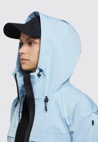 Veste imperméable bleu clair avec capuche, accents et zips noirs, et couche de base noire à côtes. Casquette noire portée sous la capuche.